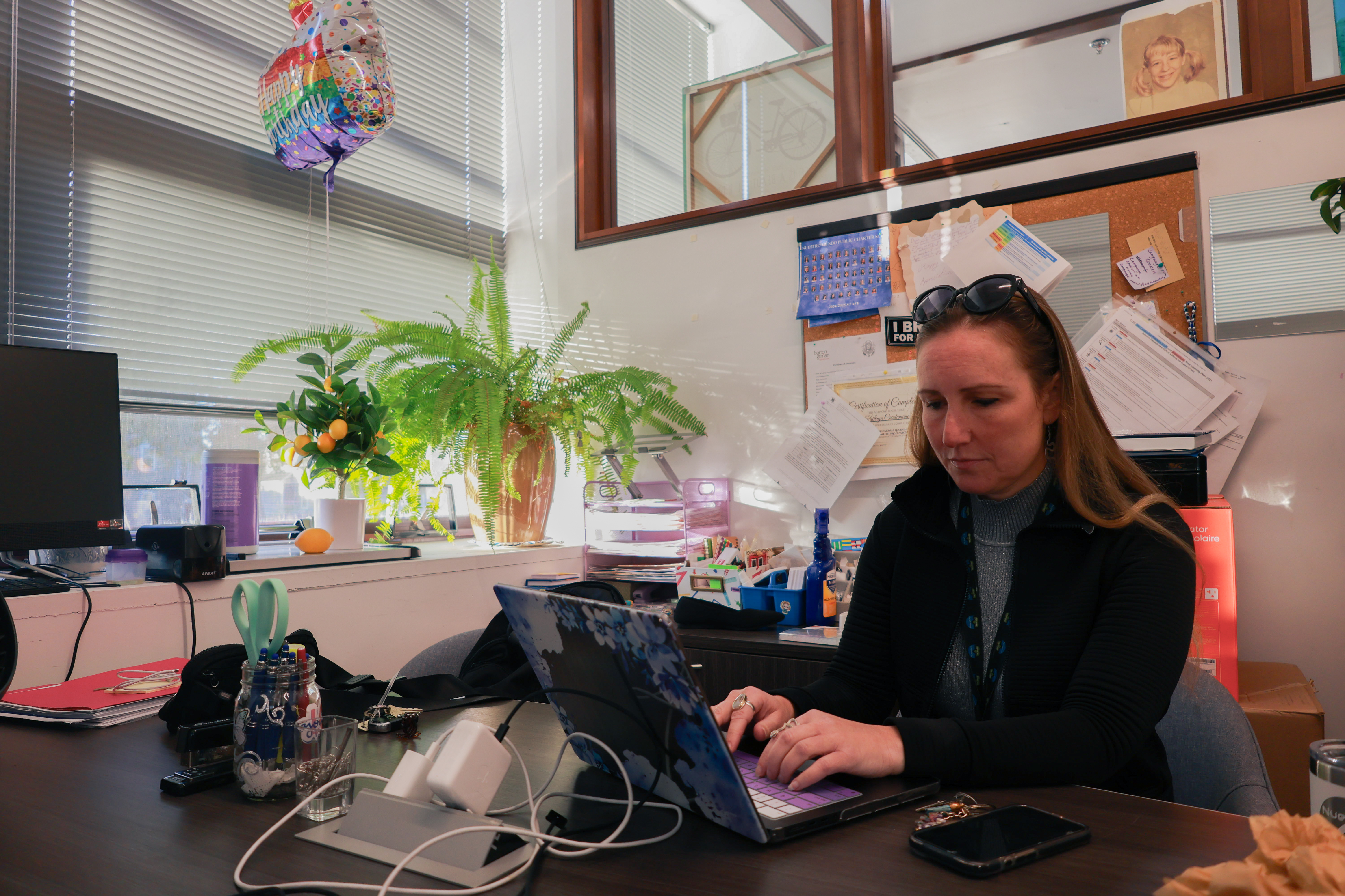 Photo of Katie Cardamone typing on her laptop in an office lit with bright morning light and decorated with two windowsill plants.